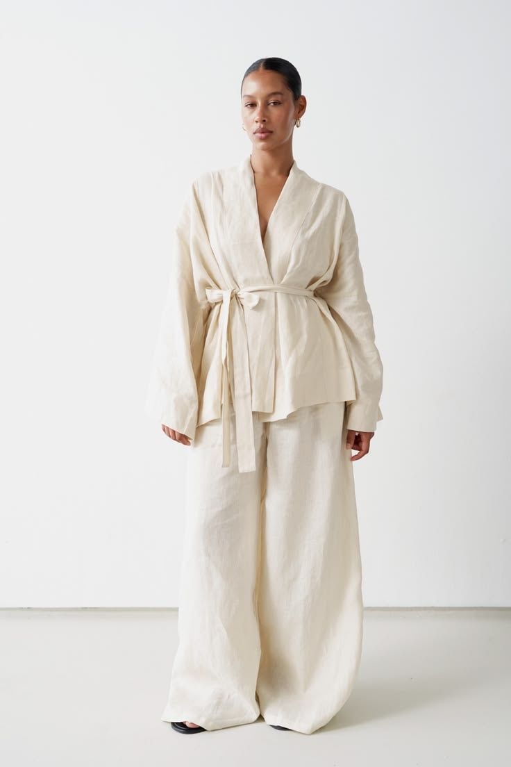 Woman in a cream belted linen jacket and wide trousers against a white studio wall