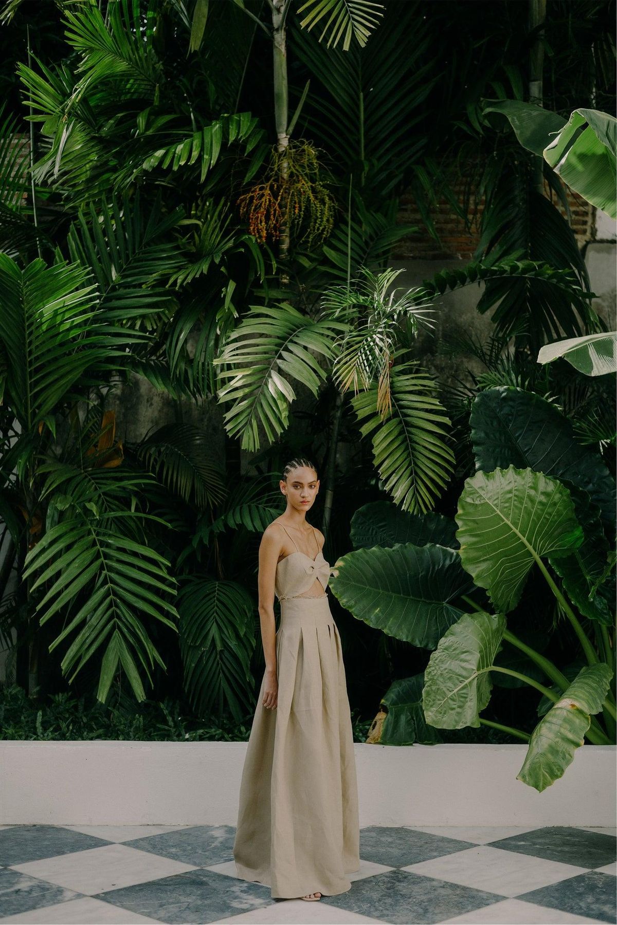 Woman in a strapless sand linen dress standing on a checkered floor before a wall of tropical leaves
