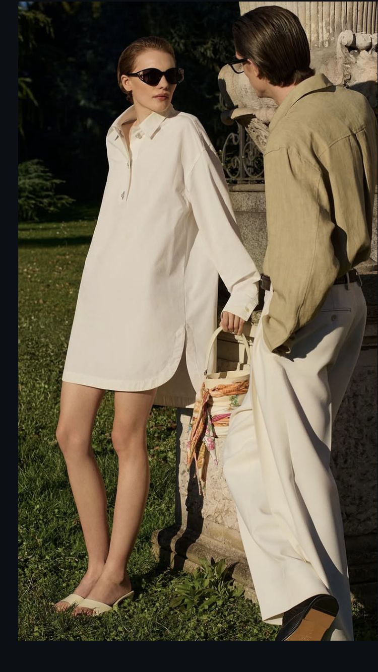Man in an olive linen shirt and cream trousers in a sunlit garden, beside a woman in a white shirt dress