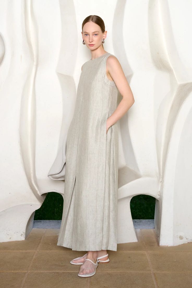 Woman in a long grey-green linen dress in front of a sculpted white wall